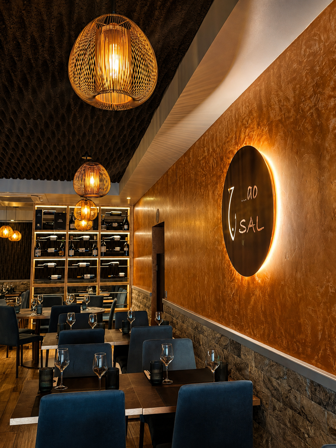 Sala de jantar com mesas postas, cadeiras azuis, parede dourada e cave de vinhos iluminada