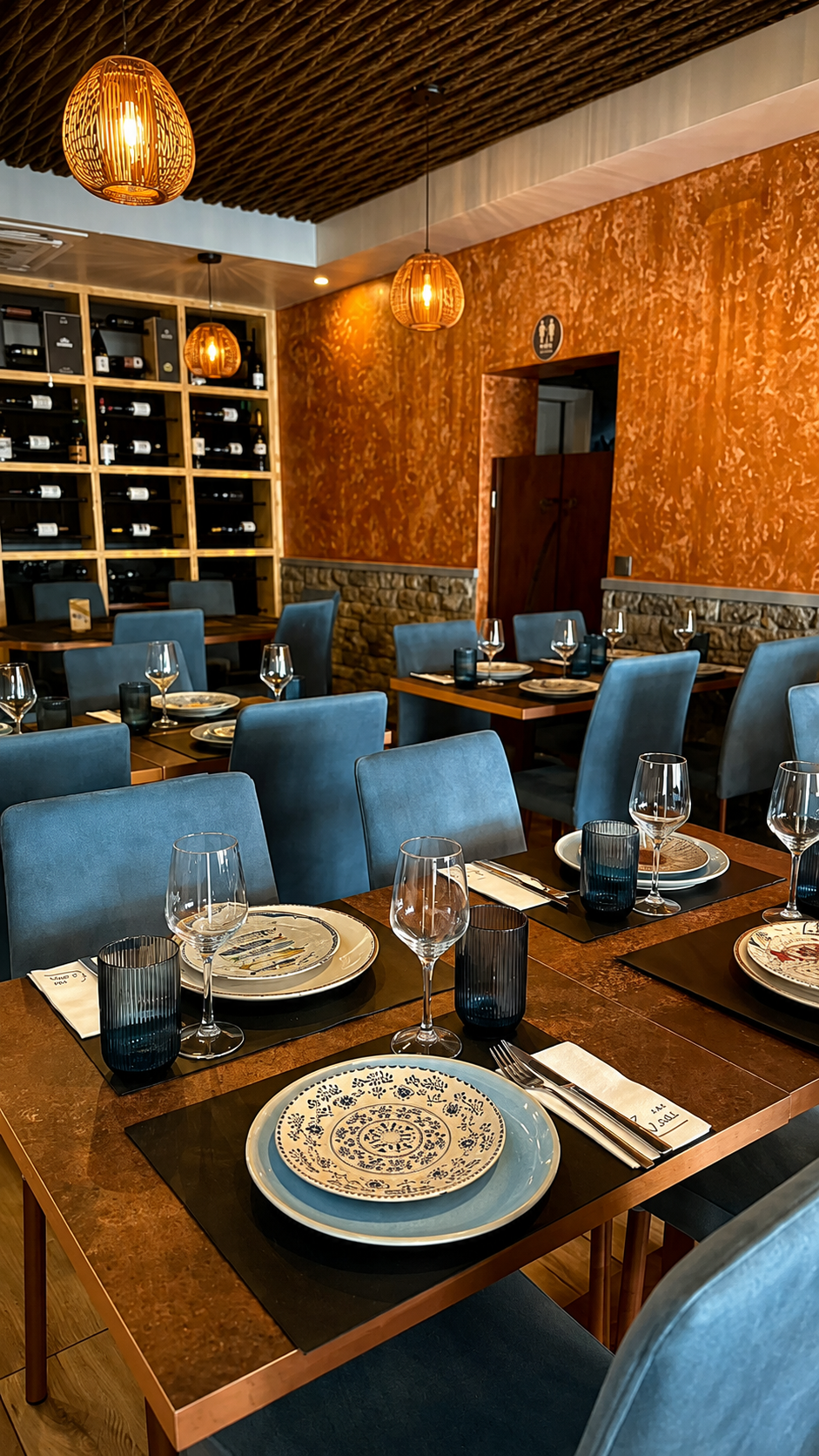 Sala de jantar com mesas postas, cave de vinhos iluminada e luminárias de vime